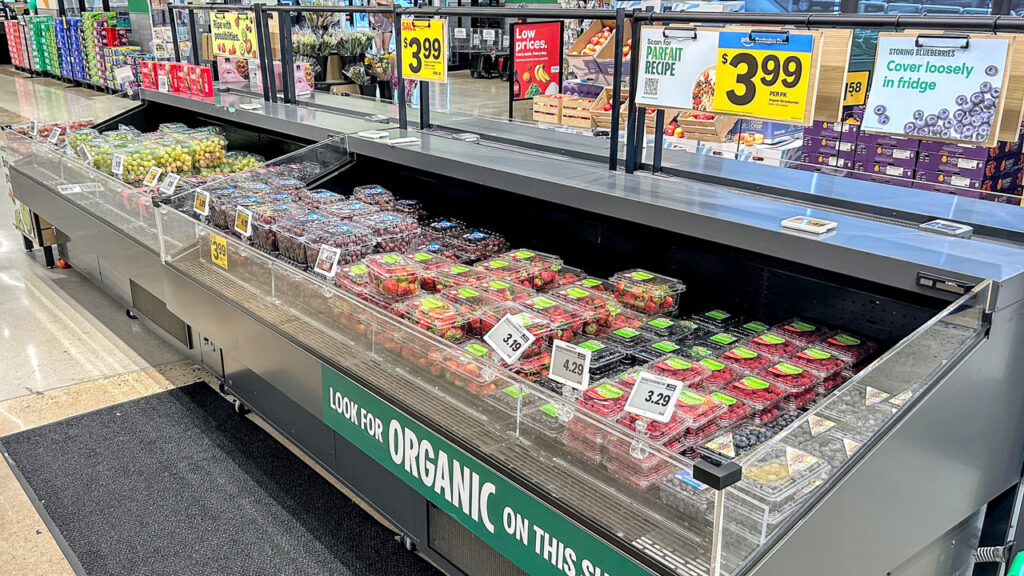 A refrigerated floor case showcases packaged berries and grapes, with paper signage above enhancing the electronic display below.